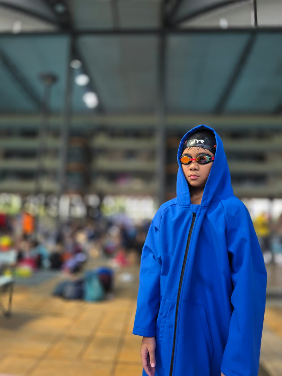 Evan in a blue swim parka, cap and goggles at a swimming venue.